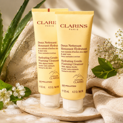 Two 125ml tubes of 2x Clarins Hydrating Gentle Foaming Cleanser are positioned upright on a stone surface, accompanied by white flowers, lush greenery, and a wooden tray, all highlighted by soft natural lighting.