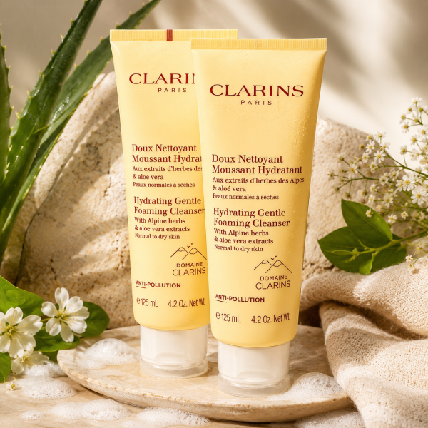 Two 125ml tubes of 2x Clarins Hydrating Gentle Foaming Cleanser are positioned upright on a stone surface, accompanied by white flowers, lush greenery, and a wooden tray, all highlighted by soft natural lighting.