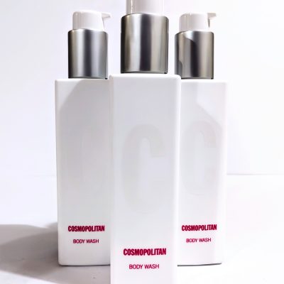 A white, rectangular bottle labeled "Cosmopolitan Body Wash" with a pump dispenser, standing on a smooth surface. Three bottles are arranged closely together.