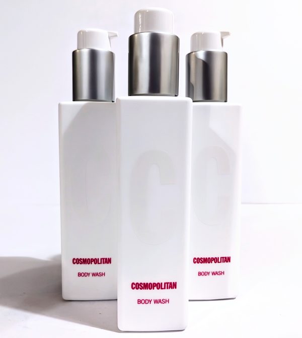 A white, rectangular bottle labeled "Cosmopolitan Body Wash" with a pump dispenser, standing on a smooth surface. Three bottles are arranged closely together.