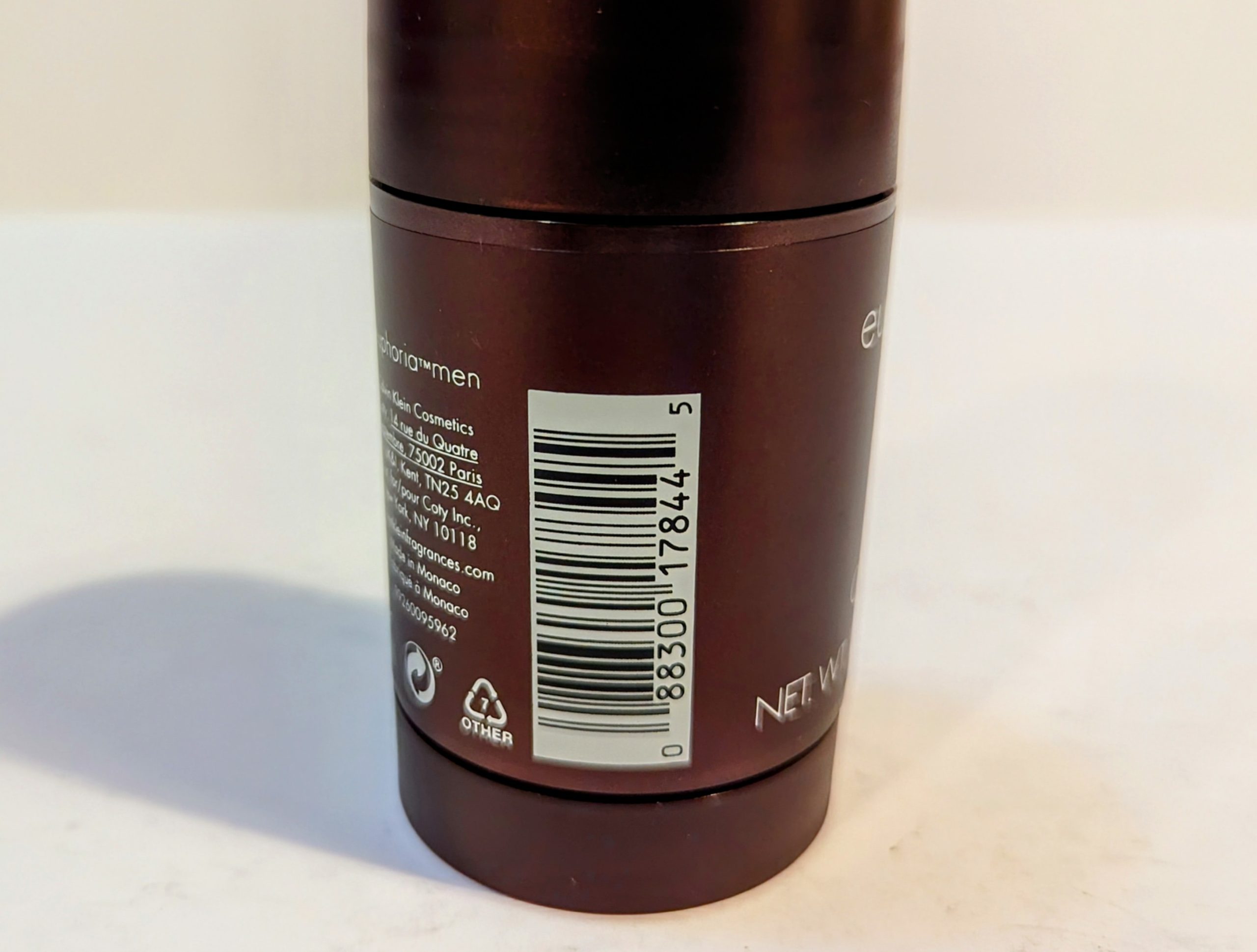 A close-up of the back of a cylindrical deodorant container displaying a barcode, recycling symbols, small text, and the phrase “NET WT 3.0 OZ.” The container is metallic brown.