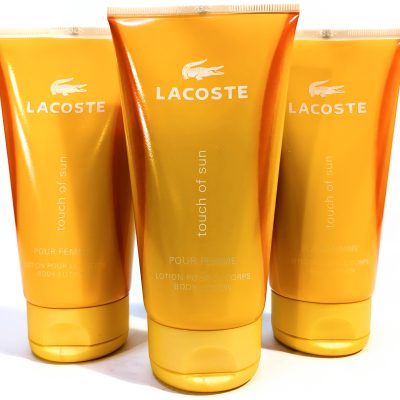 Three orange tubes of Lacoste "touch of sun" body lotion are displayed vertically, with the Lacoste logo and product name visible on each tube.