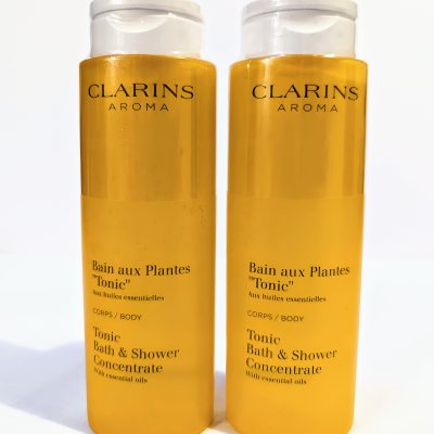 Two yellow bottles of Clarins Aroma Bath and Shower Concentrate with essential oils, labeled "Bain aux Plantes 'Tonic'" for body usage, each with a clear flip-top cap.