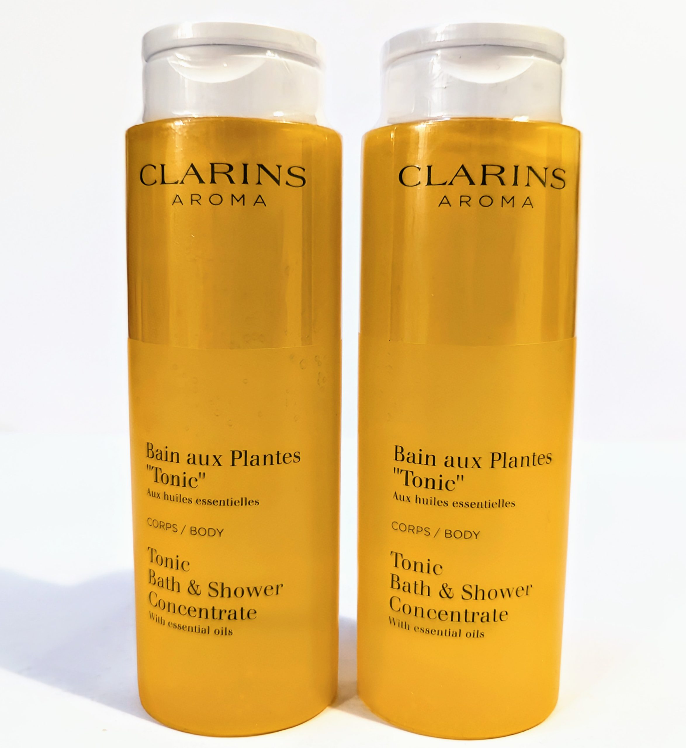 Two yellow bottles of Clarins Aroma Bath and Shower Concentrate with essential oils, labeled “Bain aux Plantes ‘Tonic'” for body usage, each with a clear flip-top cap.