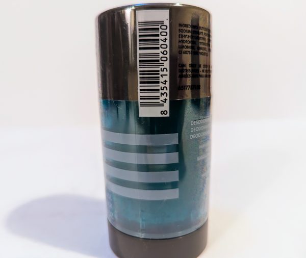 A cylindrical deodorant stick with a blue body and dark gray cap, displaying a barcode and ingredient list on the clear plastic wrapper. The label features three white horizontal lines.