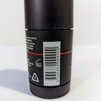 A close-up of a barcode on a cylindrical container with a black label and white text. Recycling symbols and a product longevity icon marked as "18M" are visible below the barcode.