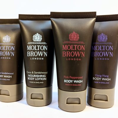 Four tubes of Molton Brown body care products are displayed. The products include White Sandalwood Body Wash, Coco & Sandalwood Nourishing Body Lotion, Pink Pepperpod Body Wash, and Ylang-Ylang Body Wash.