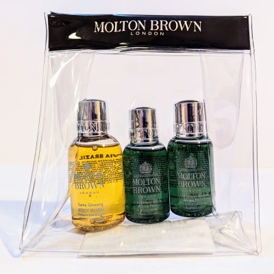 A clear plastic bag containing three Molton Brown London travel-sized bottles: one yellow Suma Ginseng body wash and two green products, likely body lotions or similar items.