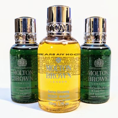 Three Molton Brown bottles: two small, green bottles flanking a larger, yellow bottle. All have mirrored silver caps and detailed labels.