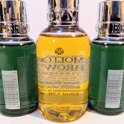 Three Molton Brown bottles with chrome caps; two green bottles flank a central yellow bottle, each displaying barcodes on their back labels.