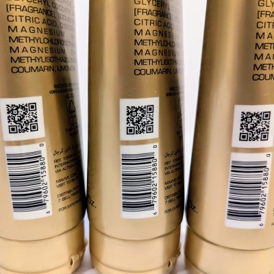 Three gold-colored product bottles are lined up with QR codes and ingredient lists visible on their labels. Each bottle also features a barcode and product information printed in multiple languages.