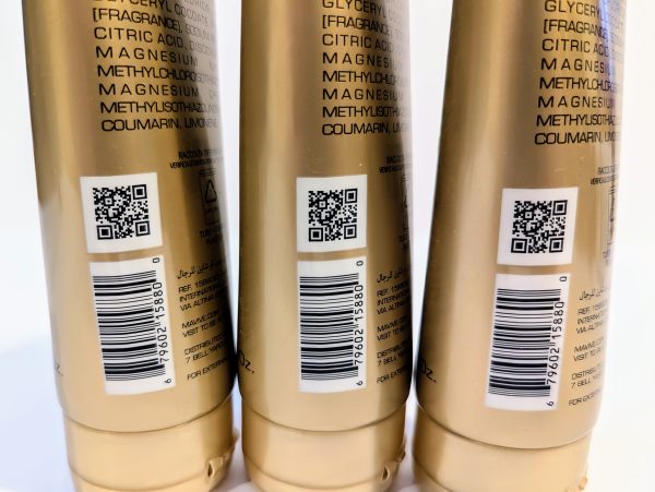 Three gold-colored product bottles are lined up with QR codes and ingredient lists visible on their labels. Each bottle also features a barcode and product information printed in multiple languages.