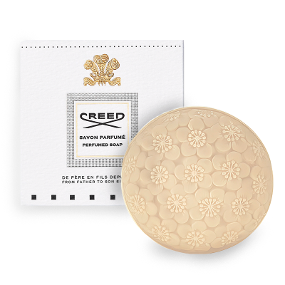 A round bar of perfumed soap embossed with floral designs is placed beside its packaging, which showcases the brand "Creed" and reads "Savon Parfumé".