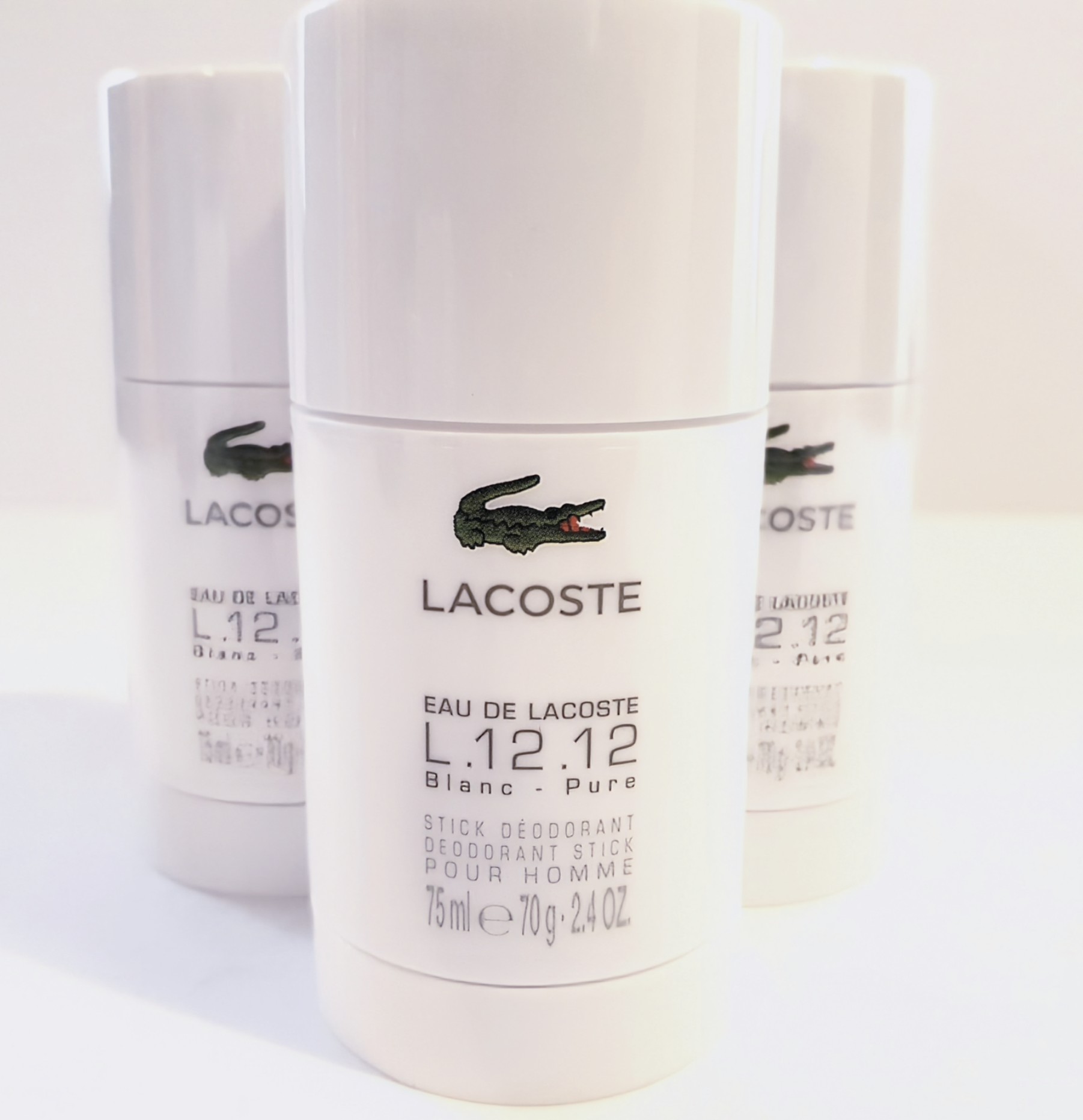 Three white Lacoste L.12.12 Blanc Pure deodorant stick containers are displayed, each holding 75 ml (2.4 oz). The containers have a green crocodile logo and product details written in black text.