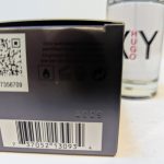 A close-up of a product box showing a QR code, text, symbols, a bar code, and a serial number. A partially visible bottle labeled "Hugo XY" is in the background.