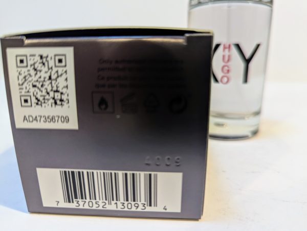 A close-up of a product box showing a QR code, text, symbols, a bar code, and a serial number. A partially visible bottle labeled "Hugo XY" is in the background.