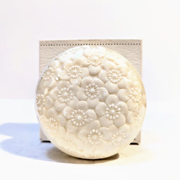 A round, white soap with intricate floral carvings is placed in front of a white box on a plain background.