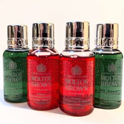 Four small bottles of Molton Brown shower gel in various colors (red and green) are arranged closely together on a light background.