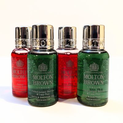 Four Molton Brown bath and shower gel bottles with shiny silver caps are shown. Two bottles are green and labeled "Fabled Juniper Berries & Lapp Pine," and the other two are red.