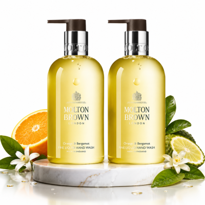 Two bottles of 2x Molton Brown Orange & Bergamot Hand Wash 300ml are shown on a marble platform with orange slices, bergamot, green leaves, and white flowers.