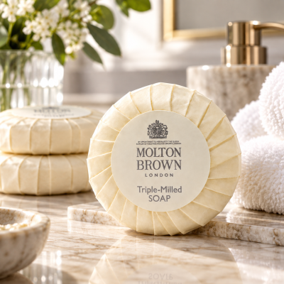 A round 3x Molton Brown Triple-Milled Milk Soap 25g Minatures rests on a marble counter with towels, stacked soaps, and a soap dish in the background.