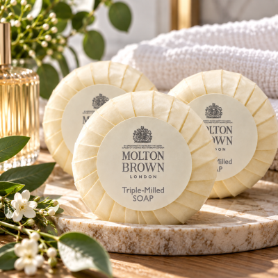 Three 3x Molton Brown Triple-Milled Milk Soap 25g Minatures are arranged on a table with greenery, flowers, a towel, and a glass perfume bottle in the background.