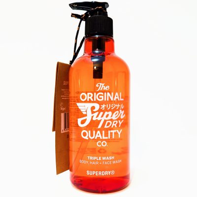 A transparent orange bottle of Superdry triple wash for body, hair, and face with a black pump and a brown tag attached.