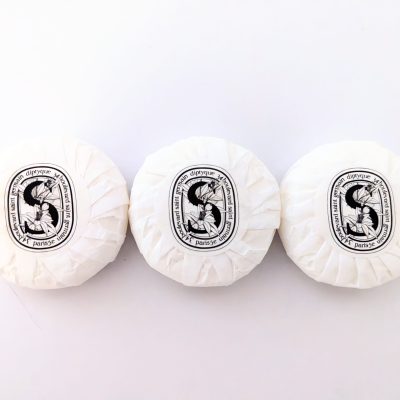 Three round Diptyque Paris soaps wrapped in white paper, each with a black and white label, arranged side by side on a plain white surface.