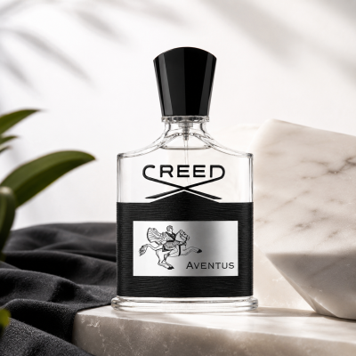 A clear glass bottle of Creed Aventus Eau de Parfum EDP 50ml Spray for Men with a black cap, displayed on a marble slab beside a green plant and a black cloth, under soft, neutral lighting.