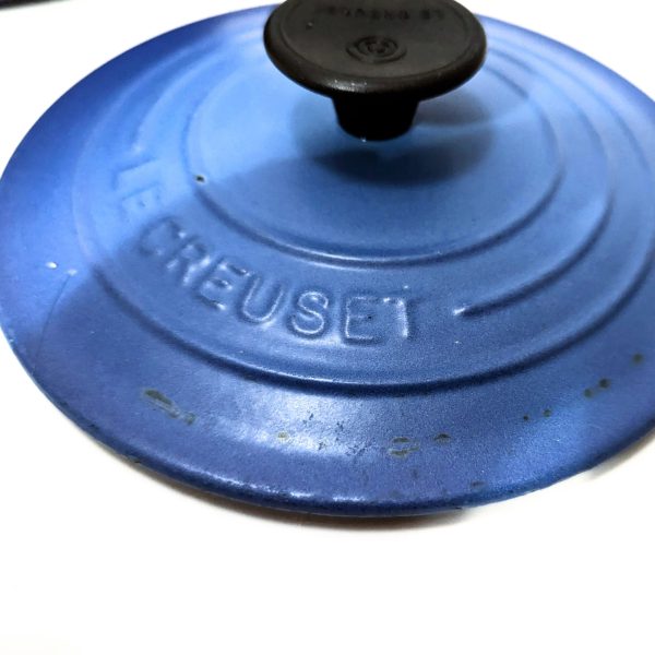Close-up of a blue Le Creuset cast iron pot lid with a black handle, showing the brand name embossed on the surface.