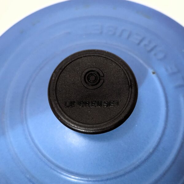 Close-up of the black knob on a blue Le Creuset cookware lid, with the brand name "Le Creuset" embossed on the knob and lid.