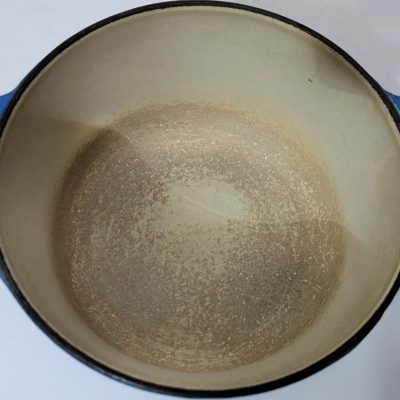 A round, blue-handled enameled cast iron pot with a worn, discolored interior sits on a white surface.
