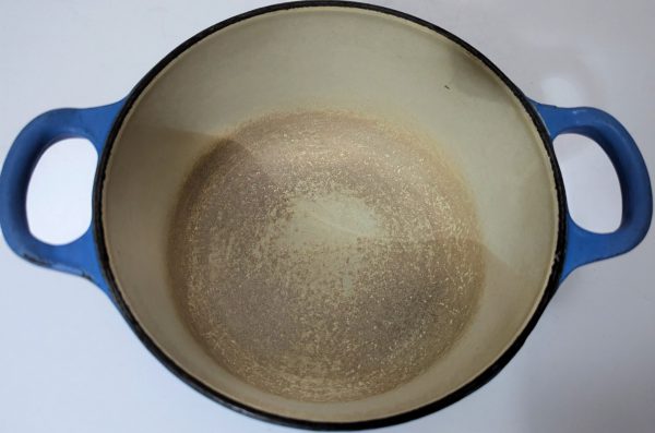 A round, blue-handled enameled cast iron pot with a worn, discolored interior sits on a white surface.