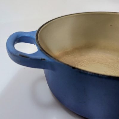 A close-up of a blue enameled cast iron pot with a beige interior, showing the handle and part of the rim.