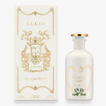 The Gucci The Alchemist's Garden Love At Your Darkest EDP Spray (100ml Unisex) is displayed in a white bottle with gold accents beside its matching decorative box adorned with gold detailing and text.