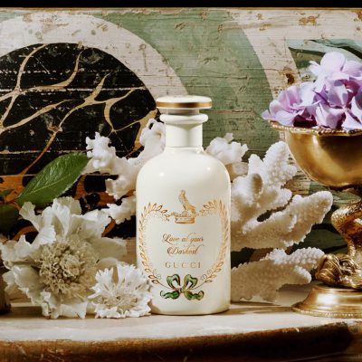 A 100ml bottle of Gucci The Alchemist's Garden Love At Your Darkest EDP Spray is displayed among white flowers, coral pieces, leaves, and a gold cup with purple flowers against a decorative background.
