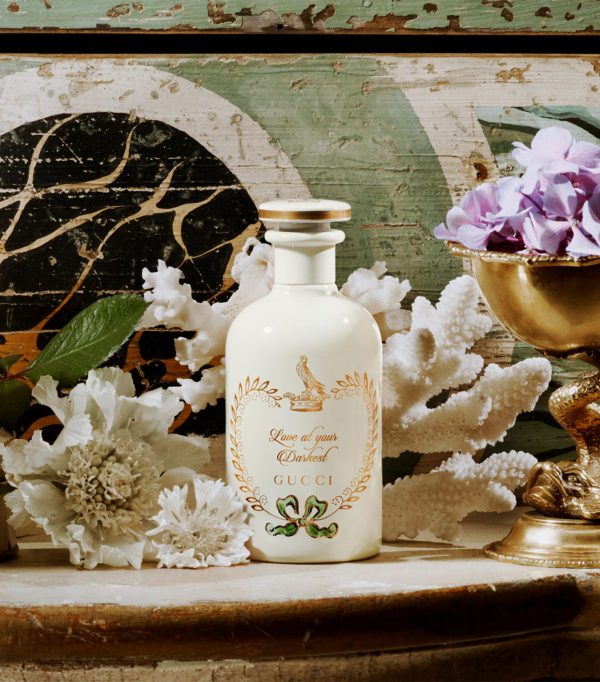A 100ml bottle of Gucci The Alchemist's Garden Love At Your Darkest EDP Spray is displayed among white flowers, coral pieces, leaves, and a gold cup with purple flowers against a decorative background.