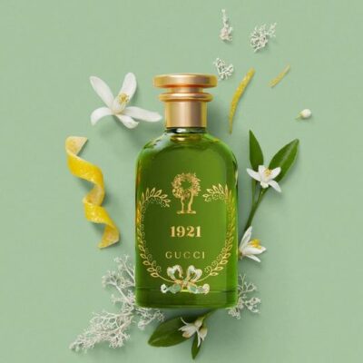 A green bottle of Gucci 1921 Eau de Parfum Spray, Unisex, 100ml sits among white flowers, green leaves, and a yellow citrus peel on a light green background.
