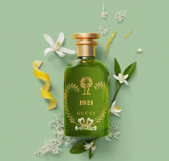 A green bottle of Gucci 1921 Eau de Parfum Spray, Unisex, 100ml sits among white flowers, green leaves, and a yellow citrus peel on a light green background.