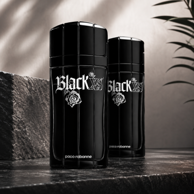 Two bottles of Paco Rabanne Black XS EDT Spray for Men, 100ml, are displayed on a dark stone surface with a plant in the background.
