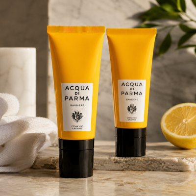 Two tubes of Acqua di Parma Barbiere Multi Action Face Cream 50ml are displayed on a marble surface with a towel, lemon half, and green leaves in the background.