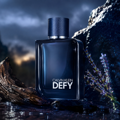 A 100ml bottle of Calvin Klein Defy Parfum Spray rests on a wet surface beside wood and lavender sprigs, with a cloudy sky behind.