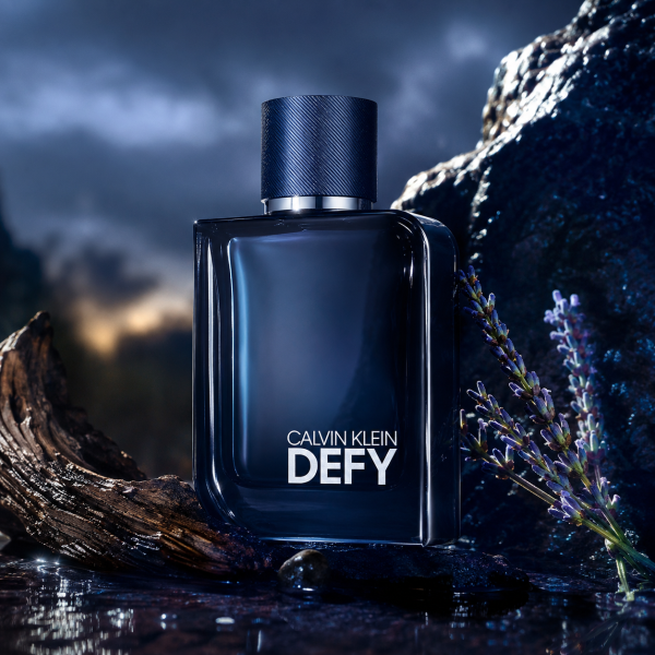 A 100ml bottle of Calvin Klein Defy Parfum Spray rests on a wet surface beside wood and lavender sprigs, with a cloudy sky behind.