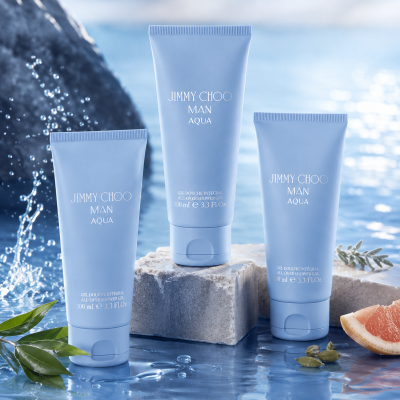 Two tubes of Jimmy Choo Man Aqua Shower Gel Body Wash for Men, 100ml each, are displayed on a wet surface with water splashes, green leaves, and a grapefruit slice nearby.
