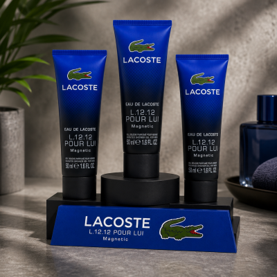 Three Lacoste L.12.12 Pour Lui Magnetic Shower Gel 50ml tubes are shown on a branded stand with a blue and green logo, placed on a stone countertop with bathroom accessories in the background.