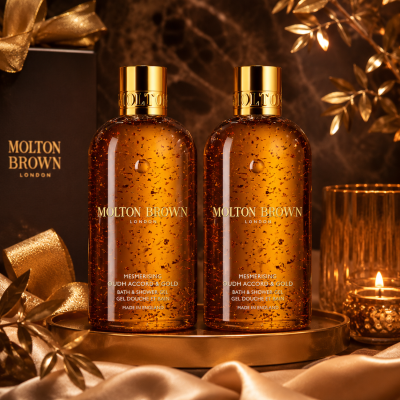 Two 300ml bottles of Molton Brown Mesmerising Oudh Accord & Gold Bath & Shower Gel are displayed on a gold surface, with a candle, ribbon, and gift box in the background.