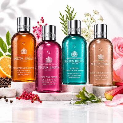 The Molton Brown Woody & Floral Body Care Shower Gel Collection (4x 100ml) is shown on a marble surface, with each bottle in various colors, surrounded by flowers, fruits, and spices.