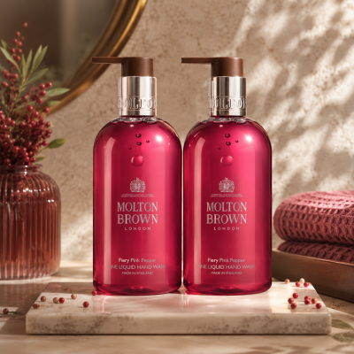 Two bottles of Molton Brown Fiery Pink Pepper Hand Wash (unisex, 300ml each) sit on a marble tray, accompanied by a pink towel, pink berries, and a glass vase in the background.