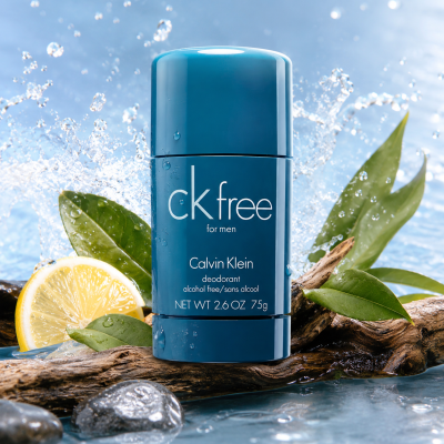 Three CK Free Deodorant Sticks for Men (75ml) in blue packaging are arranged on wet wood, surrounded by a lemon slice, fresh green leaves, and water droplets in the background.