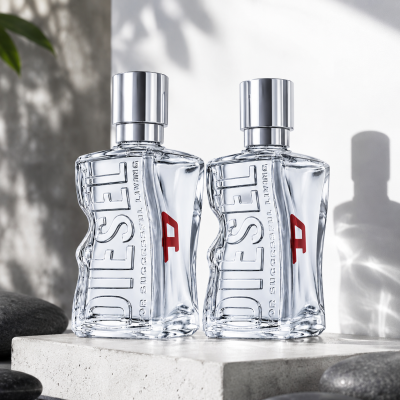 Two clear bottles of Diesel D by Diesel Eau de Toilette Spray for Men, 30ml, featuring silver caps and red accents, are placed side by side on a smooth stone surface against a white background, illuminated by soft natural light.